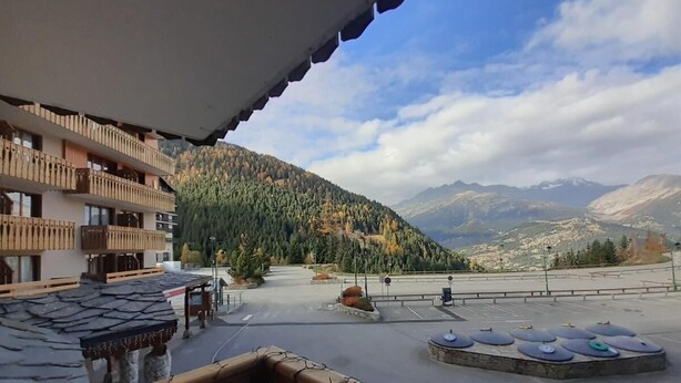 A hotel balcony with a view of a mountainous landscape, a parking area, and a pool with exercise equipment.