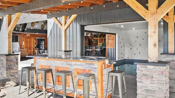 Un área de bar al aire libre con mostrador de piedra y taburetes, vigas de madera y vistas al interior.