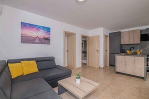 Un soggiorno moderno con un divano grigio, un tavolino da caffè con una pianta e un angolo cottura con pensili in legno e zoccolo piastrellato.