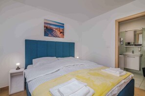 Una camera d'albergo con un letto, un comodino, un bagno con lavandino e specchio, e una scena con spiaggia raffigurata sulla parete.