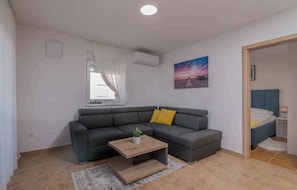 Un salon moderne avec un canapé gris, une table basse en bois et une petite plante. Il y a une fenêtre avec des rideaux blancs, un climatiseur et un tableau représentant un coucher de soleil accroché au mur.