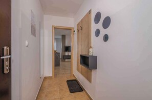 Un couloir avec une porte sur la gauche, un sol carrelé, un paillasson noir à l’entrée, un panneau en bois avec une étagère sur la droite, et un mur orné de motifs circulaires.