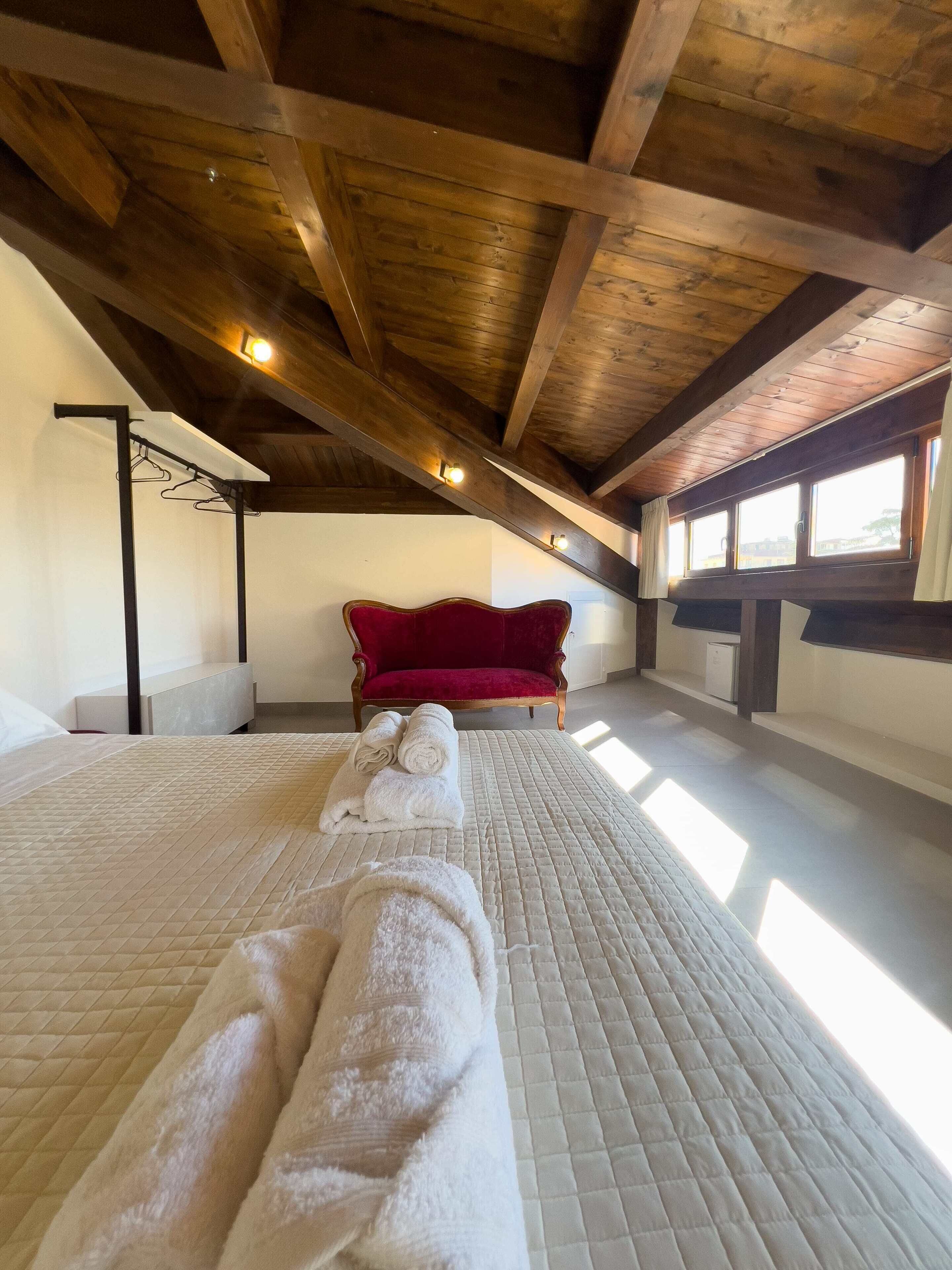 A spacious attic bedroom with a high ceiling, wooden beams, a red velvet chair, and a bed with rolled towels.