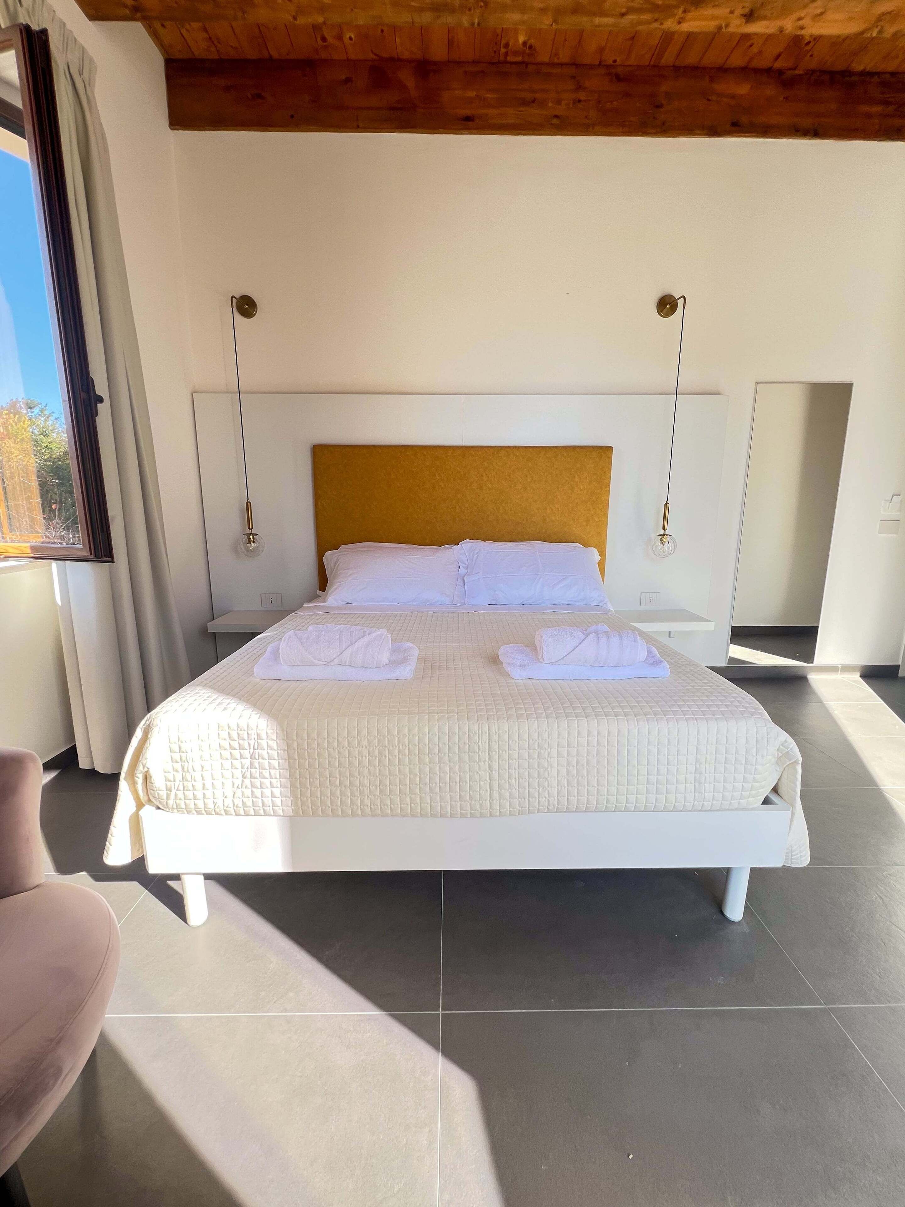 Una cama bien hecha con cabecera blanca y colchón acolchado, una mesita de noche y una ventana con cortinas.
