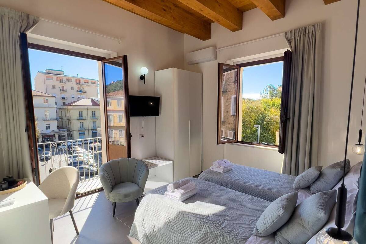 A modern bedroom with a large bed, two chairs, a TV, and a balcony with a city view.