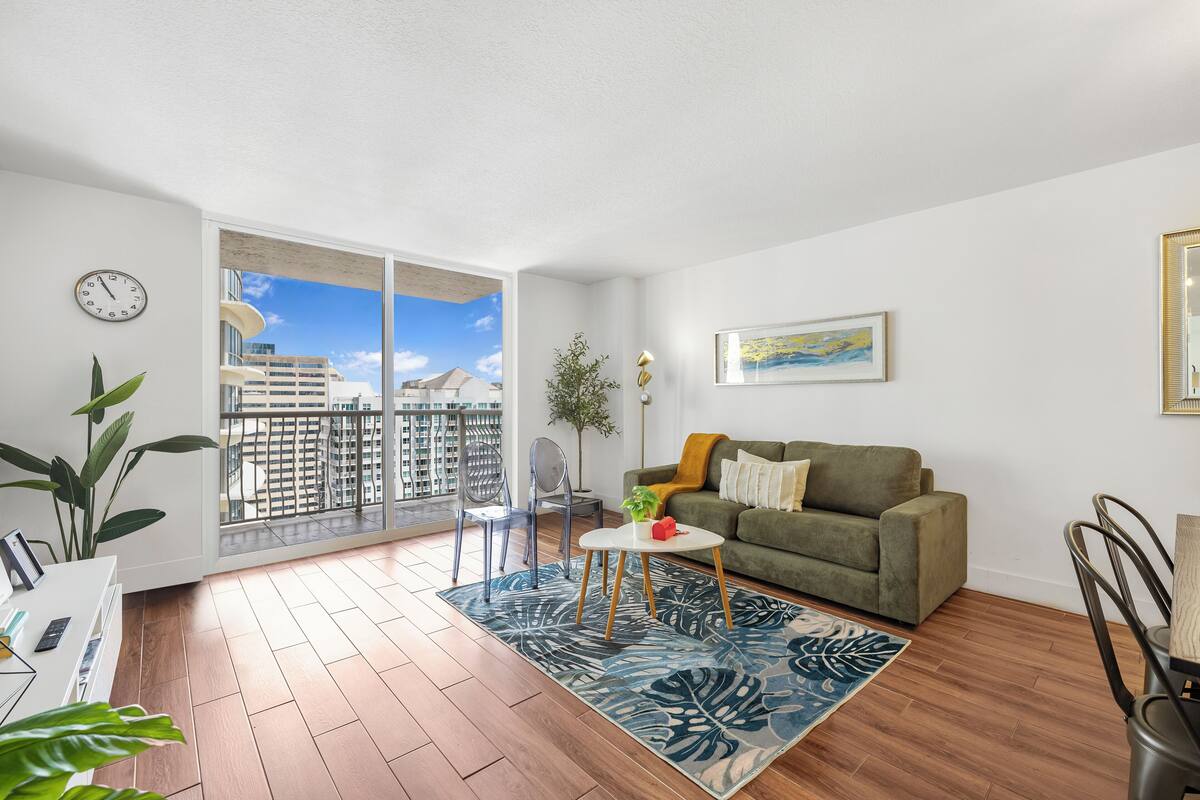 A modern living room with a wooden floor, a green sofa, a coffee table, and a view of a cityscape through large windows.