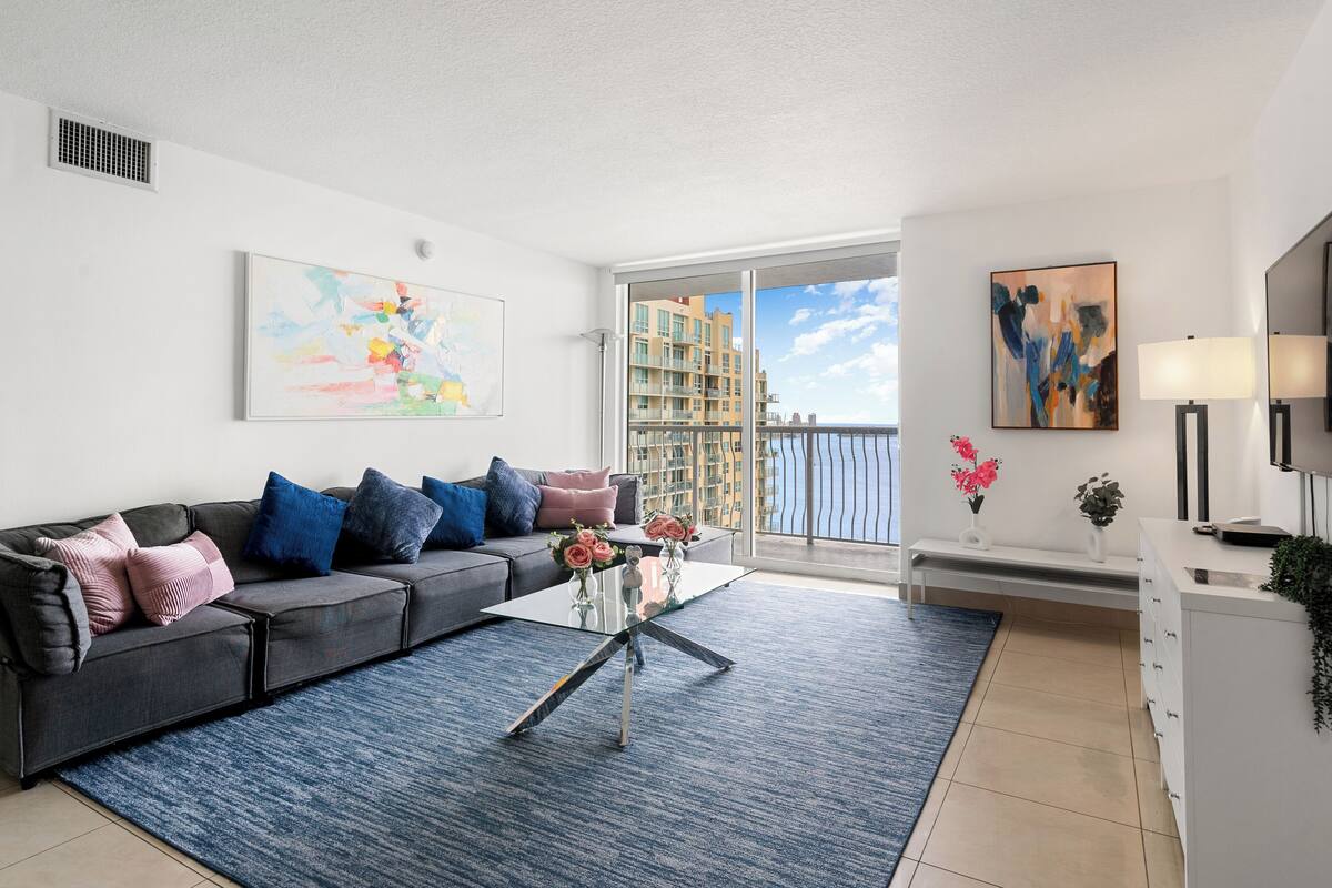 A modern living room with a grey sofa, a glass coffee table, and a view of a building through a large window.