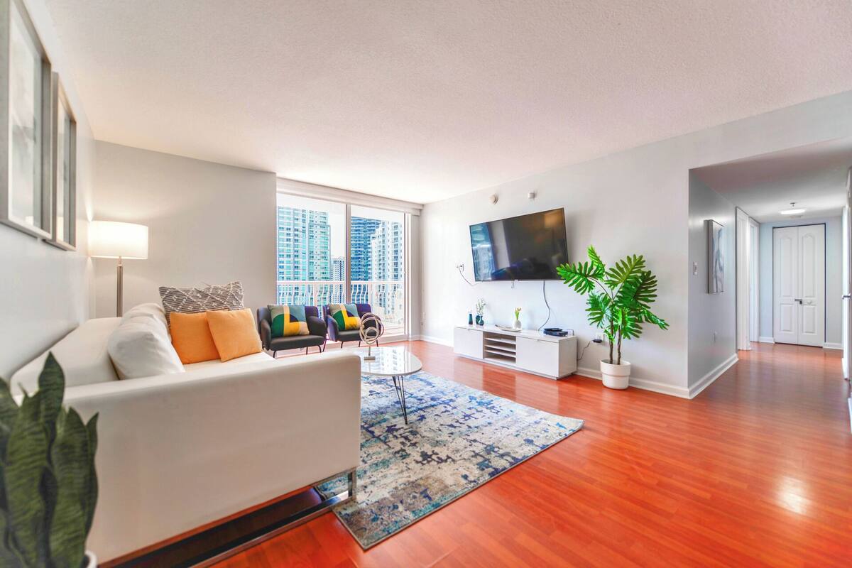 A modern living room with a white sofa, colorful cushions, a flat-screen TV, a potted plant, and a large window offering a city view.