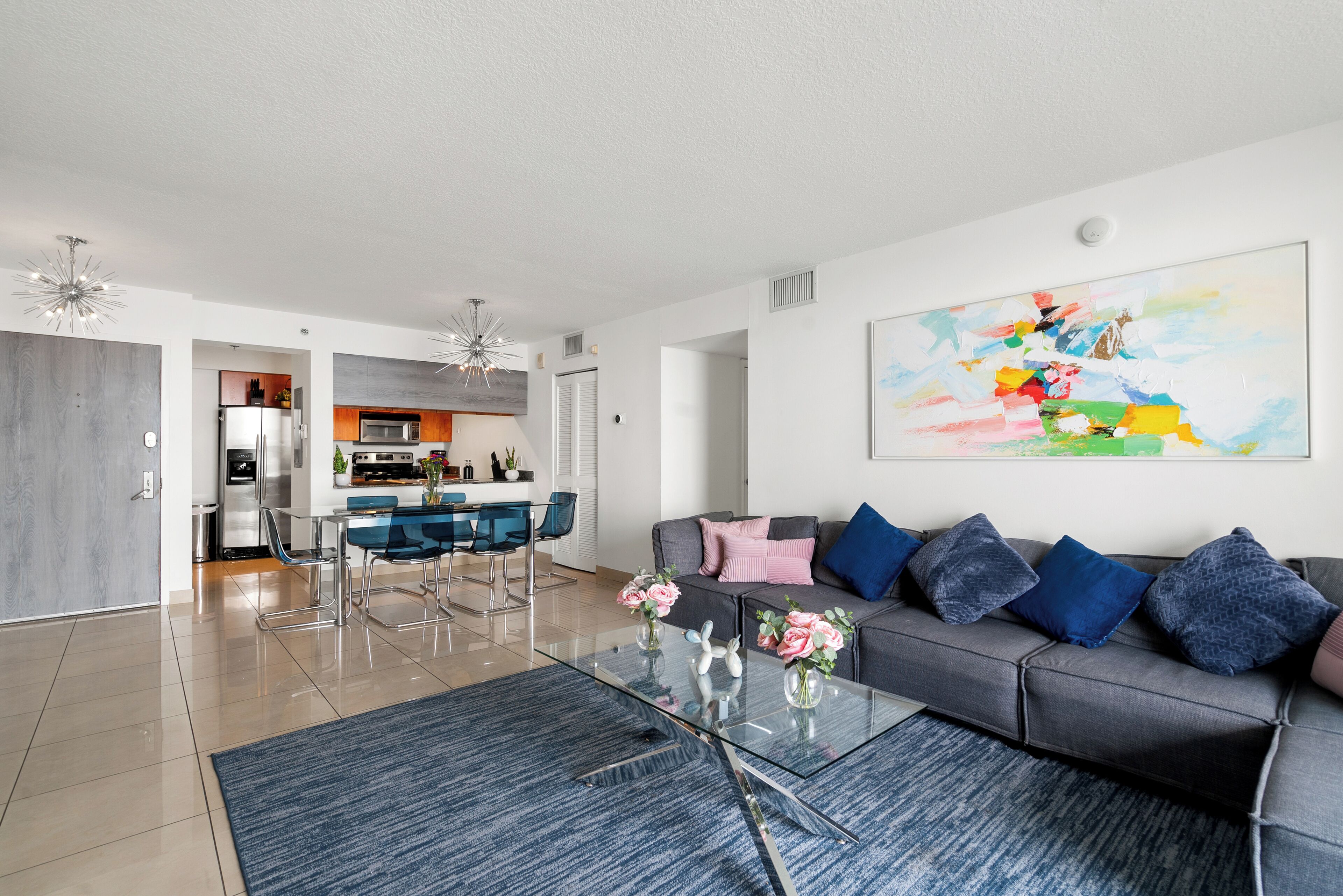 Un salon moderne avec une table basse en verre, un canapé gris agrémenté de coussins bleus et un tableau abstrait aux couleurs vives accroché au mur.