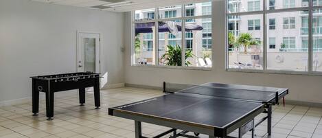 A room with a ping pong table, a foosball table, and large windows offering a view of a building and outdoor plants.