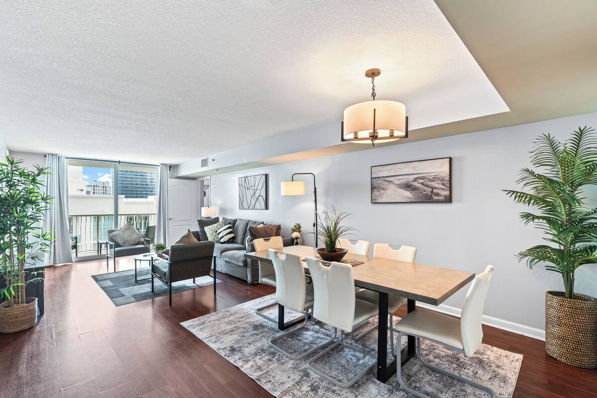 A modern living room with a dining area, large windows, and a ceiling light fixture.