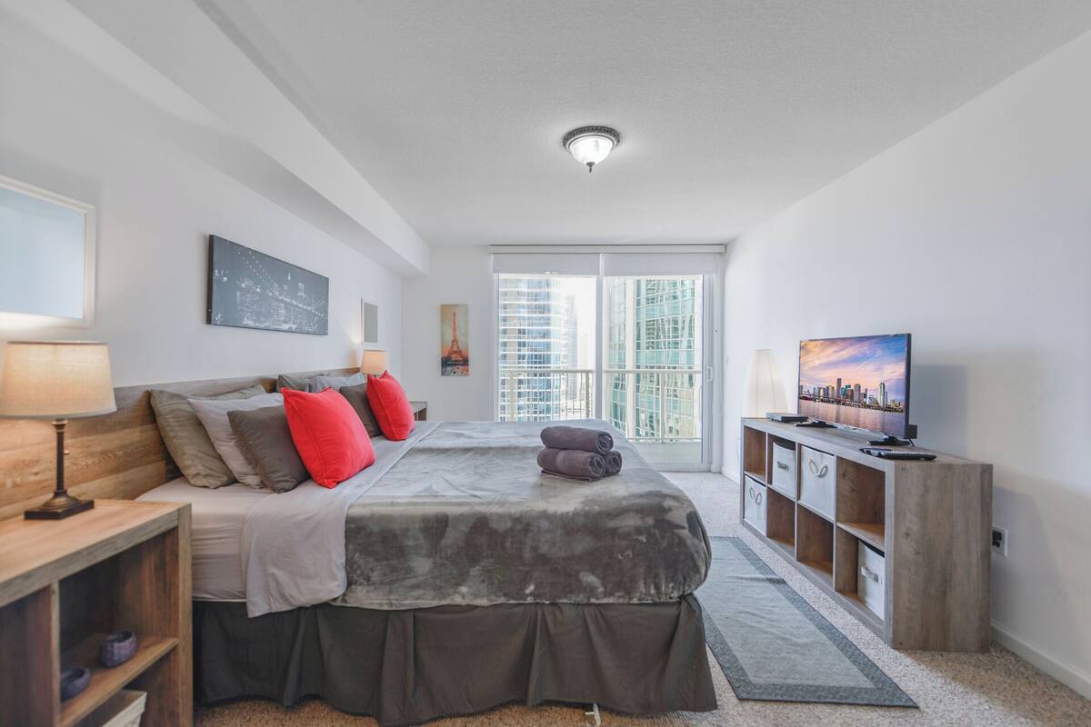 A modern bedroom with a bed, bedside tables, a TV, and a view of the city through large windows.
