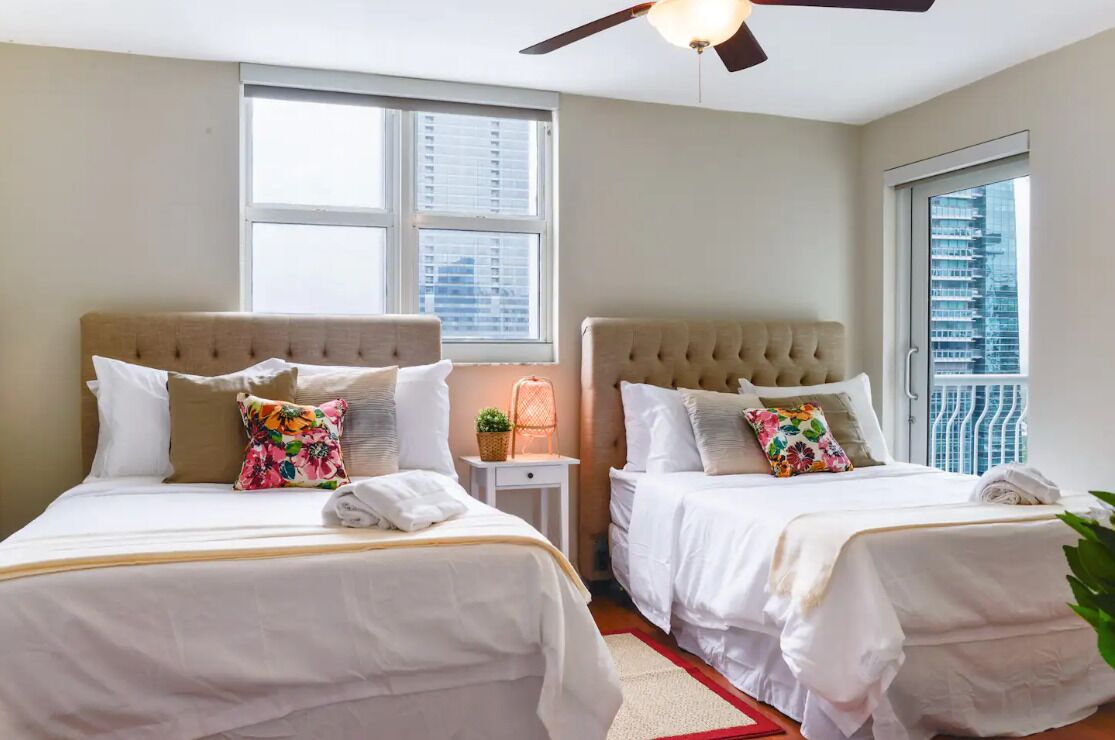 A hotel room with two beds, a ceiling fan, and large windows offering a city view.