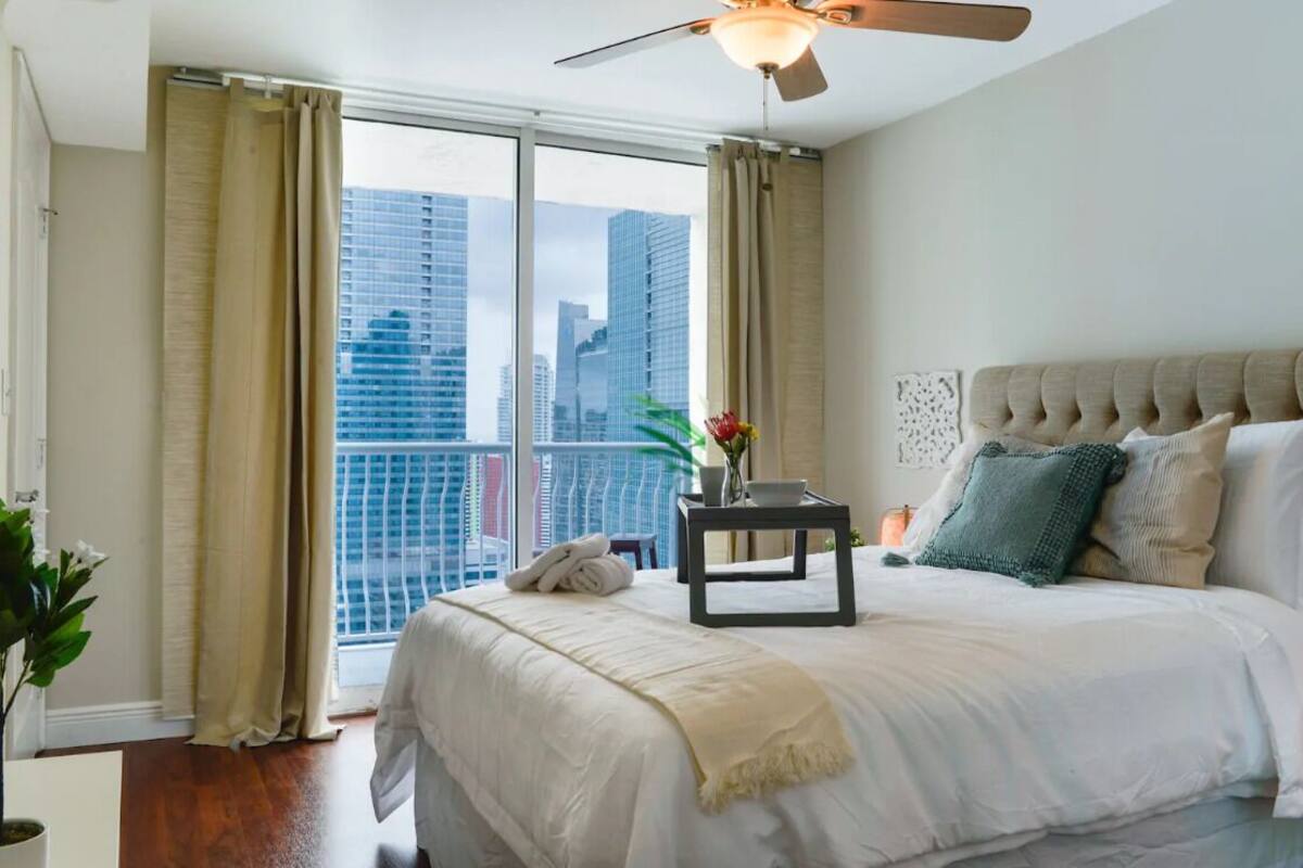 A bedroom with a large bed, a ceiling fan, and a view of a cityscape through large windows.