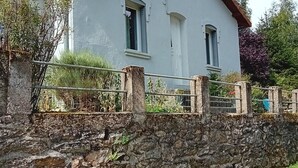 A two-story house with a stone wall in front, a metal gate, and a grassy area beside a paved path. - House and garden for 6 people near lakes and hiking trails (Luc)