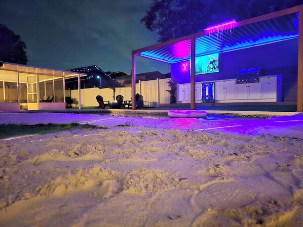 Een moderne buitenkeuken met een zwembad, overdekte zitplaatsen en een bar met neonverlichting.
