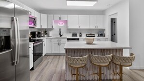 A modern kitchen with white cabinets, a stainless steel refrigerator, a built-in oven, a sink, and a bar area with stools.