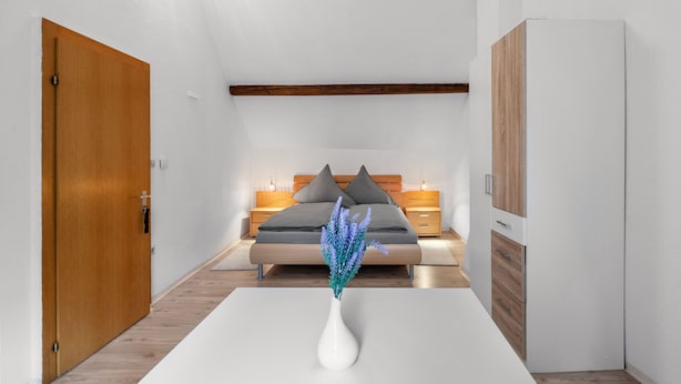 Een moderne slaapkamer met een houten deur, een witte nachtkastje met een bloemenvaas, een bed met grijze beddengoed en kussens, en een witte garderobe.
