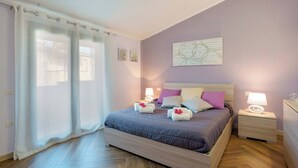 Un dormitorio con cama, dos mesitas de noche, una mesita auxiliar, una ventana con cortinas y un cuadro en la pared.