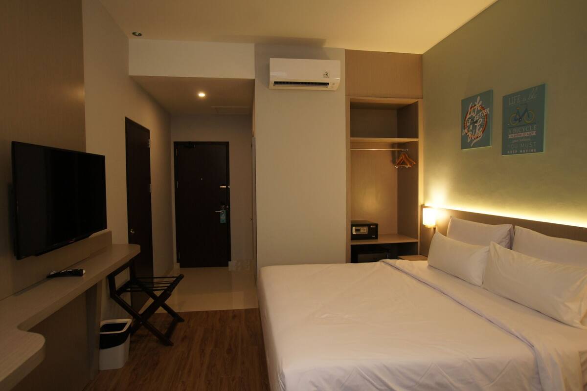 Quarto de hotel com cama, televisão, ar-condicionado, armário e parede com dois cartazes motivacionais.