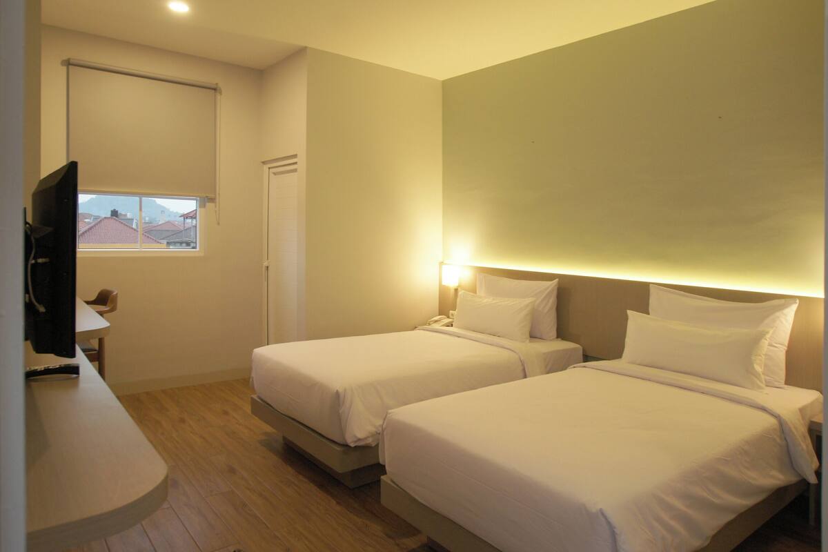 Quarto de hotel com duas camas, televisão, escrivaninha e janela com vista para edifícios.