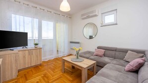Un salon moderne avec un plancher en bois, un canapé gris, une table basse en bois ornée d’un vase de fleurs, un téléviseur à écran plat posé sur un meuble en bois, et de grandes fenêtres avec des rideaux légers.