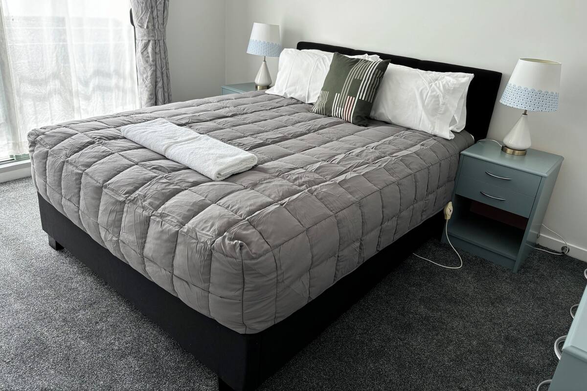 A neatly made bed with a grey quilt, two bedside lamps, and a small drawer unit beside it.