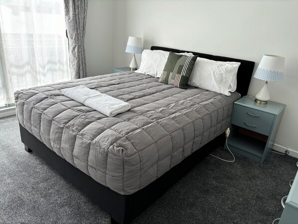 Una cama bien hecha con un edredón gris, dos lámparas de noche y una cómoda pequeña a su lado.