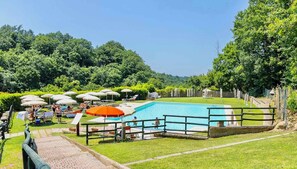 Ein Schwimmbecken, umgeben von Gras und Bäumen, mit Sonnenschirmen und Liegestühlen.