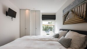 Ein Schlafzimmer mit Bett, einem an der Wand befestigten Fernseher, einem Kleiderschrank und einem Fenster mit Blick ins Freie.