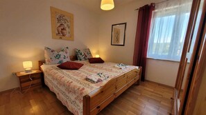 Ein Schlafzimmer mit einem hölzernen Bett, zwei Nachttischen, einer Nachttischlampe, einem Fenster mit Vorhängen und zwei an der Wand befestigten Bildern.
