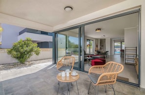 A modern outdoor patio with wicker chairs and a table, a glass door leading to an interior dining area, and a small tree in the background. - Villa with sea view and pool (Malinska)