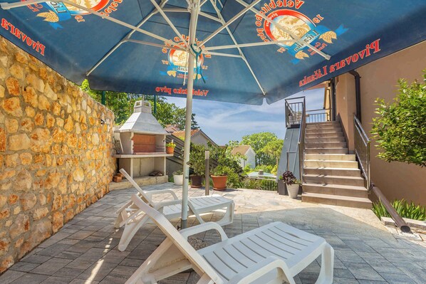 A stone wall, outdoor seating area with white chairs, a barbecue grill, and a staircase leading to a second floor.