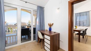 Una habitación con un escritorio de madera, una silla, una mesa de comedor con sillas, un jarrón con flores secas y vistas a través de puertas de cristal amplias.
