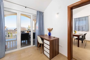 Una habitación con un escritorio de madera, una silla, una mesa de comedor con sillas, un jarrón con flores secas y vistas a través de puertas de cristal amplias.