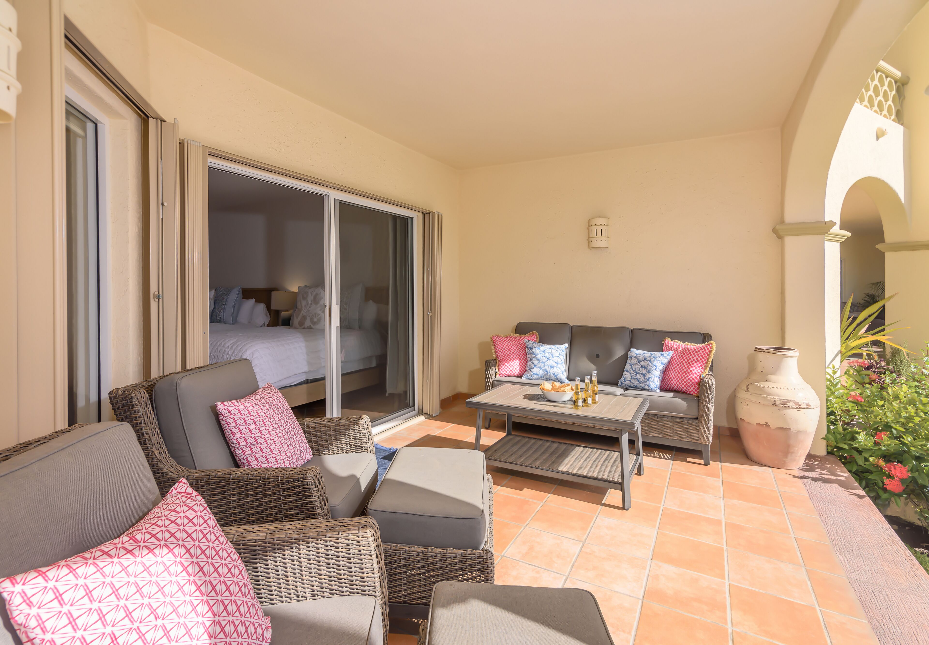A sunlit patio with wicker furniture, a sofa with patterned cushions, a coffee table, and a large vase.