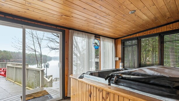 A wooden cabin with a hot tub, large windows, and a view of a lake.