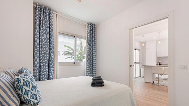 A bedroom with a bed, patterned pillows, a window with curtains, and a view of a kitchen and bathroom.
