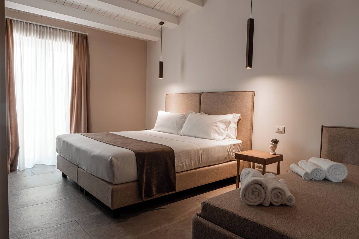 A modern hotel room with a large bed, a bedside table with towels, and a small table with a vase.