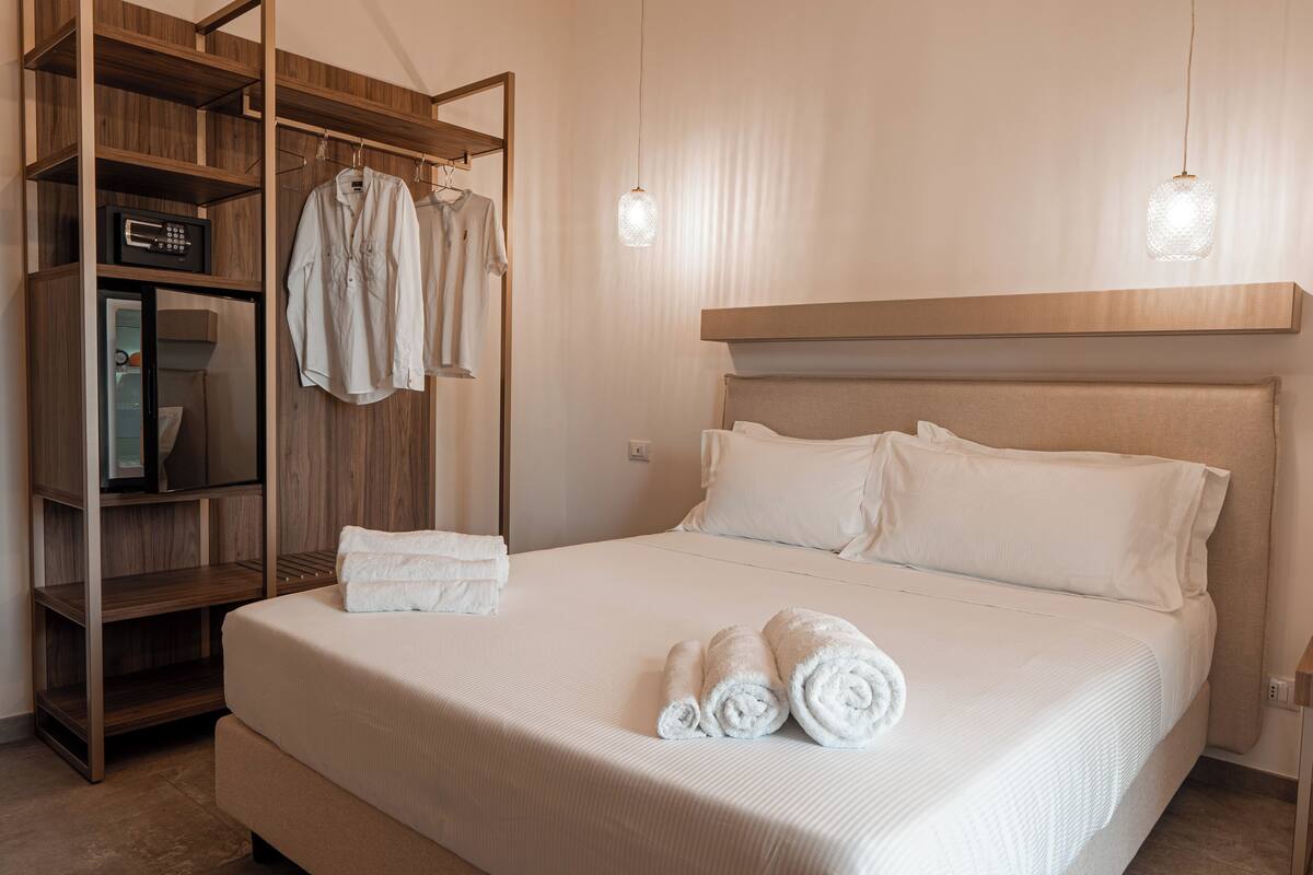 A neatly made bed with white linens and towels, a wooden wardrobe with hanging clothes, and a modern pendant light.