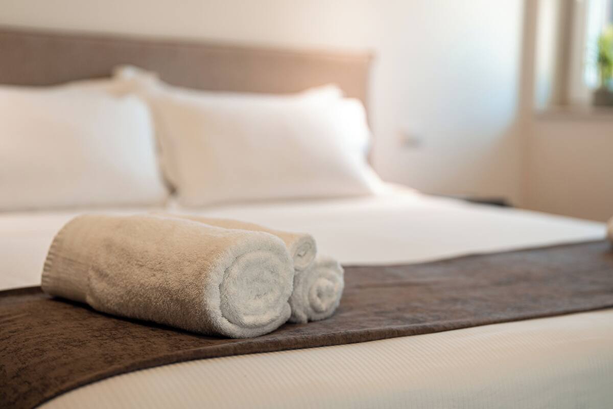 A neatly made bed with white linens and a rolled white towel on a wooden tray.