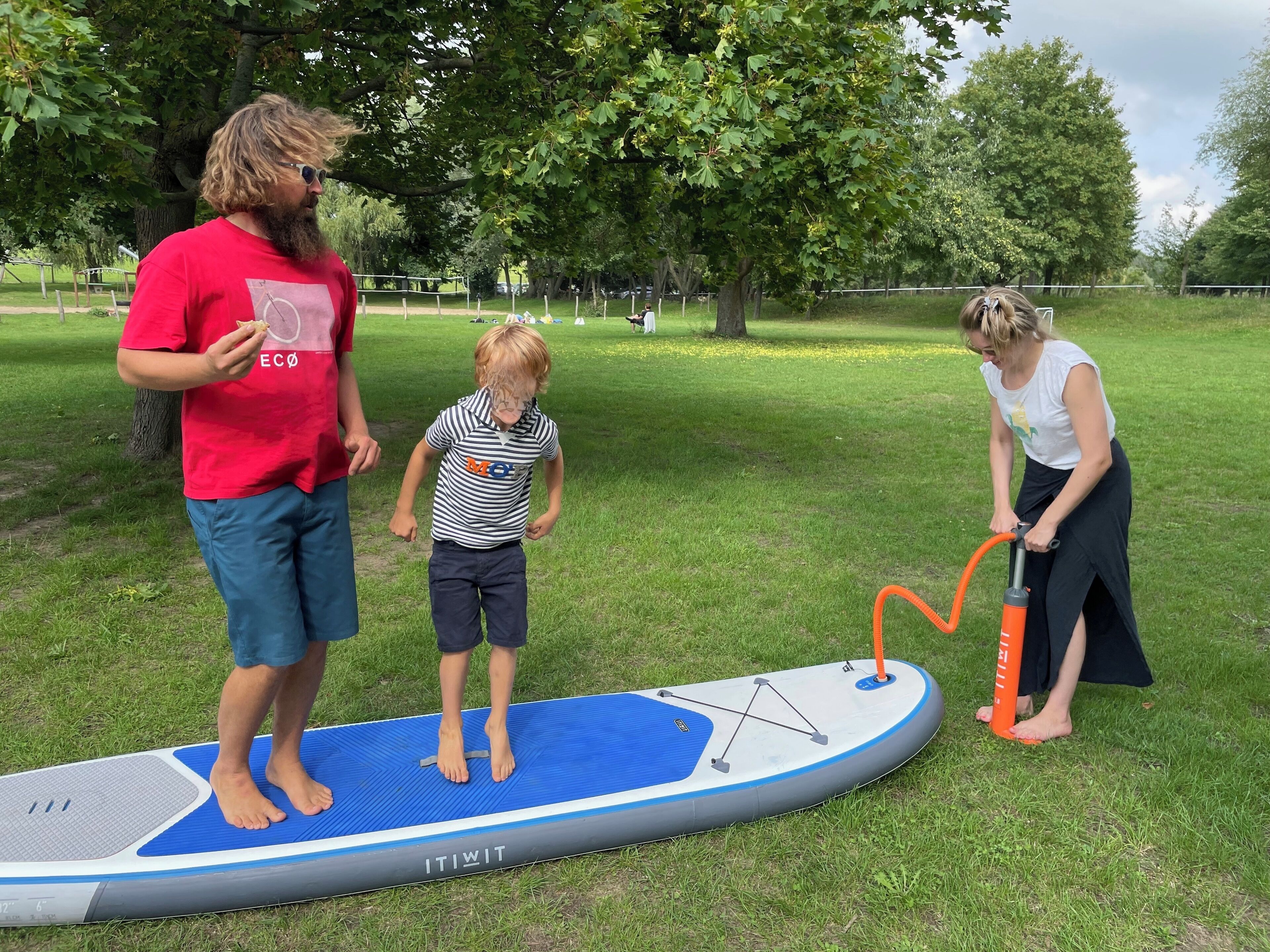 Drie mensen staan op een blauw en wit paddleboard in een parkachtig grasgebied.