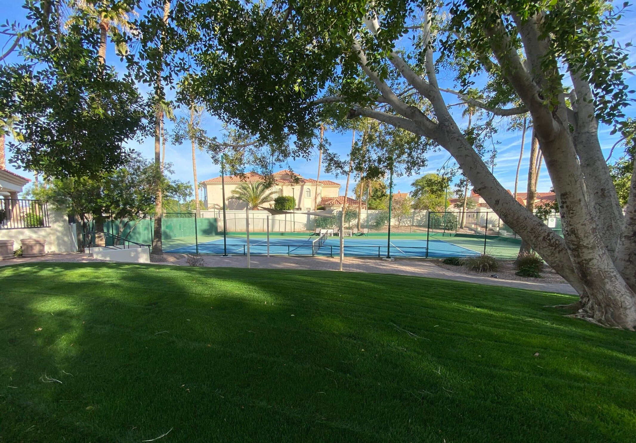 Un espace verdoyant bien entretenu, avec un court de tennis, entouré d’arbres et d’un bâtiment dont le toit est recouvert de tuiles rouges.