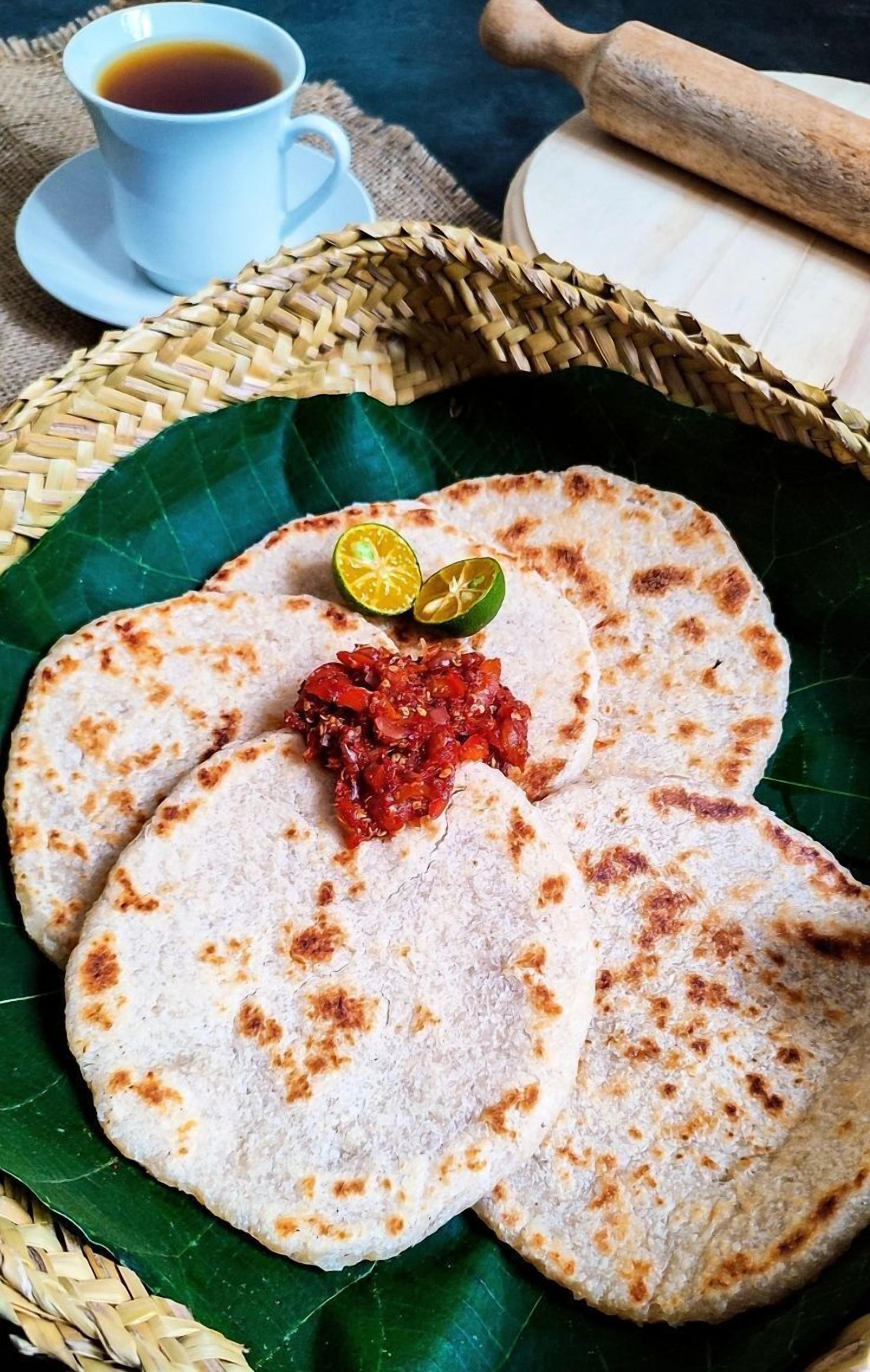 A basket of flatbreads with a red condiment, a cup of tea, and a rolling pin on a woven mat.