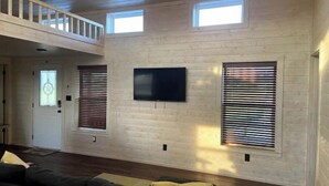 Un vaste salon doté d’un haut plafond, d’un grand téléviseur à écran plat fixé au mur et d’un escalier en bois menant à un niveau supérieur.