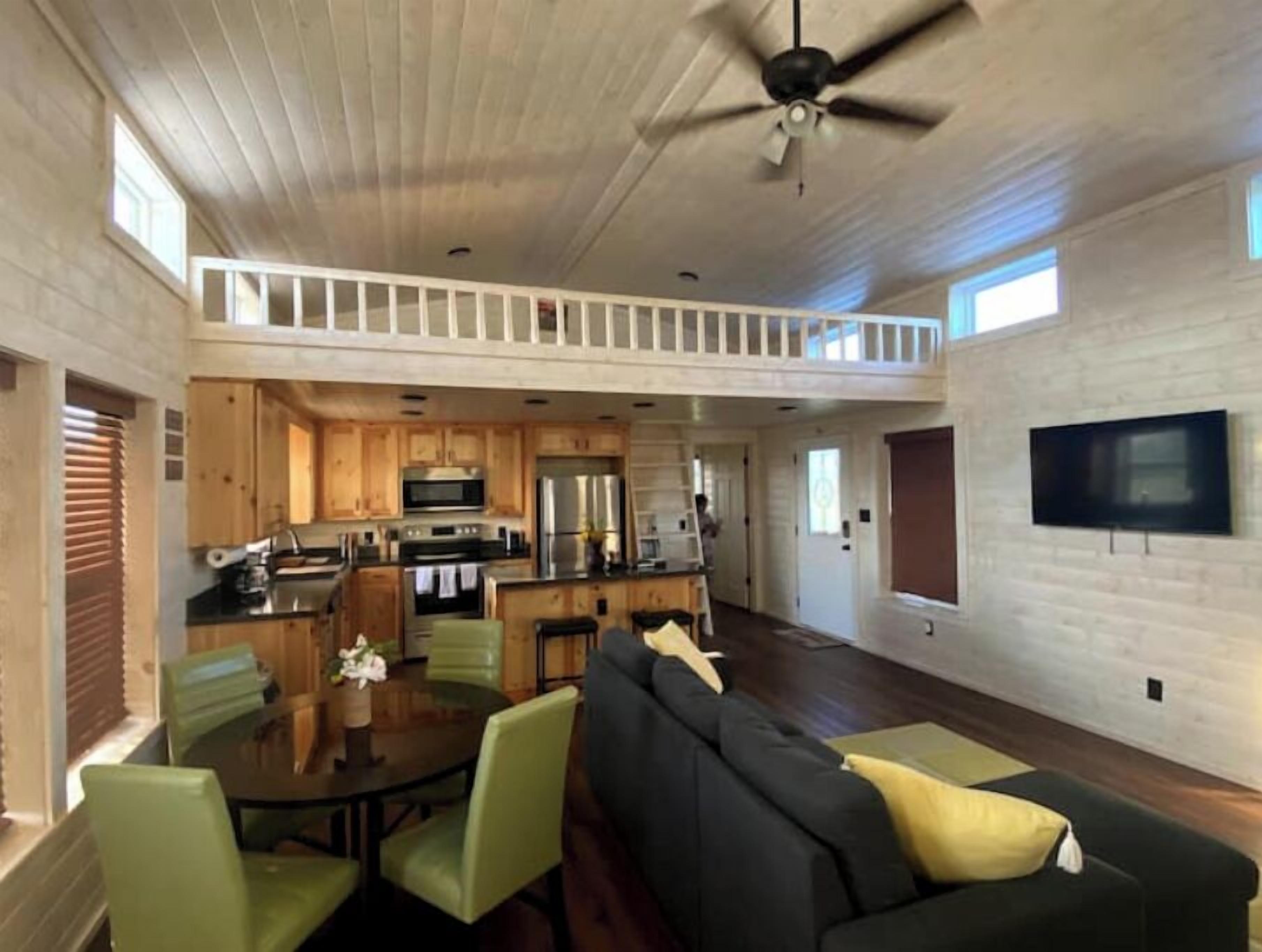 A spacious living area with a high ceiling, a dining table with chairs, a sofa, and a kitchen area with wooden cabinets and modern appliances.