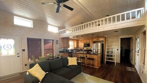 Un vaste espace de vie avec un canapé aux couleurs sombres, une cuisine dotée de meubles en bois et un ventilateur de plafond.