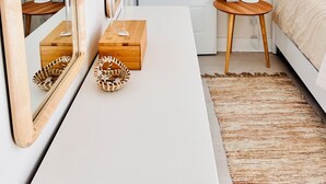 Una camera da letto con una cassettiera bianca, un comodino in legno, un letto con coperta beige, una porta bianca e un pannello decorativo in macramè a parete.
