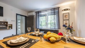 Una mesa de comedor dispuesta con platos, cubiertos y un frutero, con una ventana y cortinas al fondo.