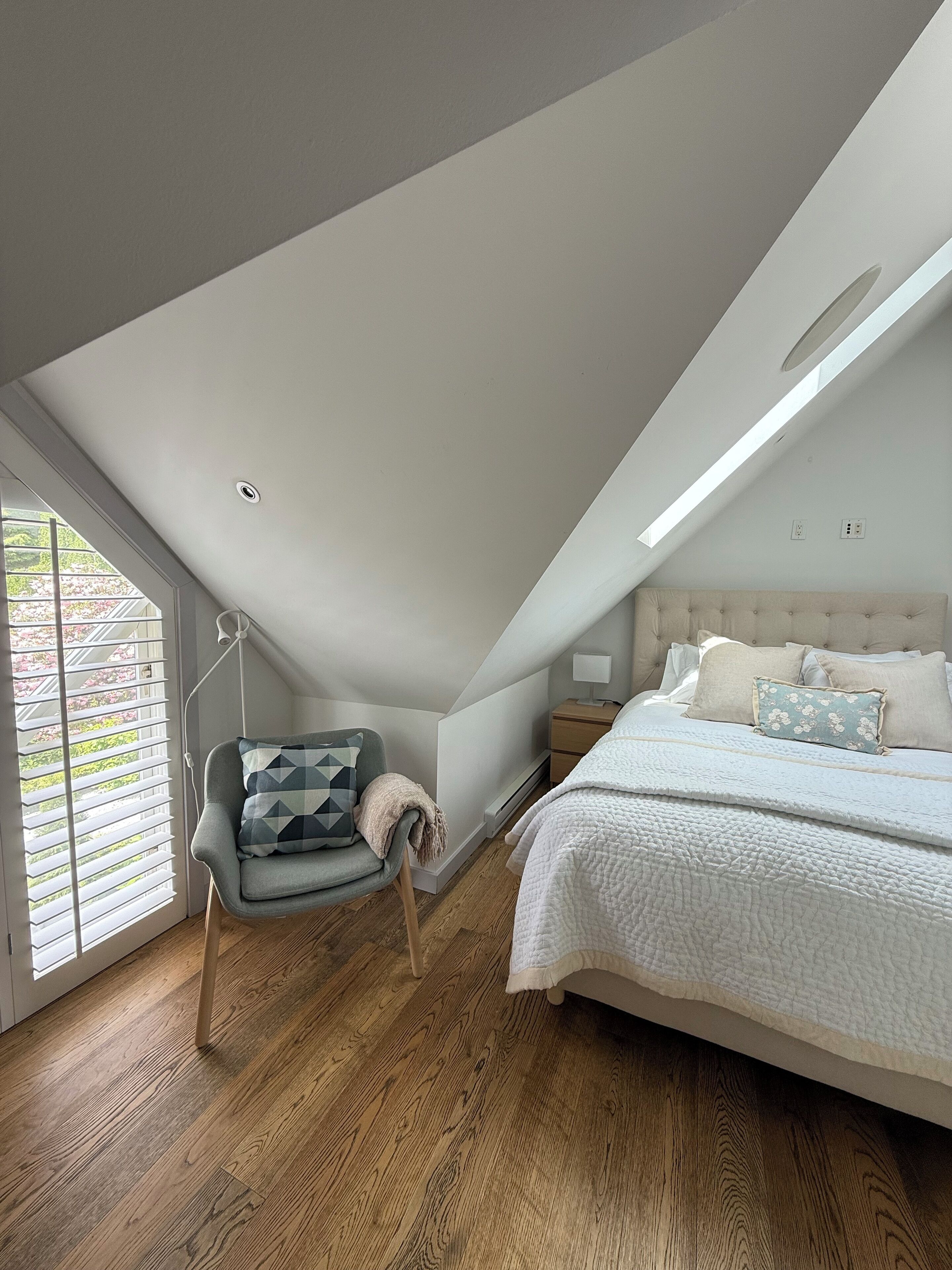 Un dormitorio con techo inclinado, una cama con ropa de cama blanca, una mesita de noche, una silla con un cojín de diseño geométrico y una ventana con persianas.